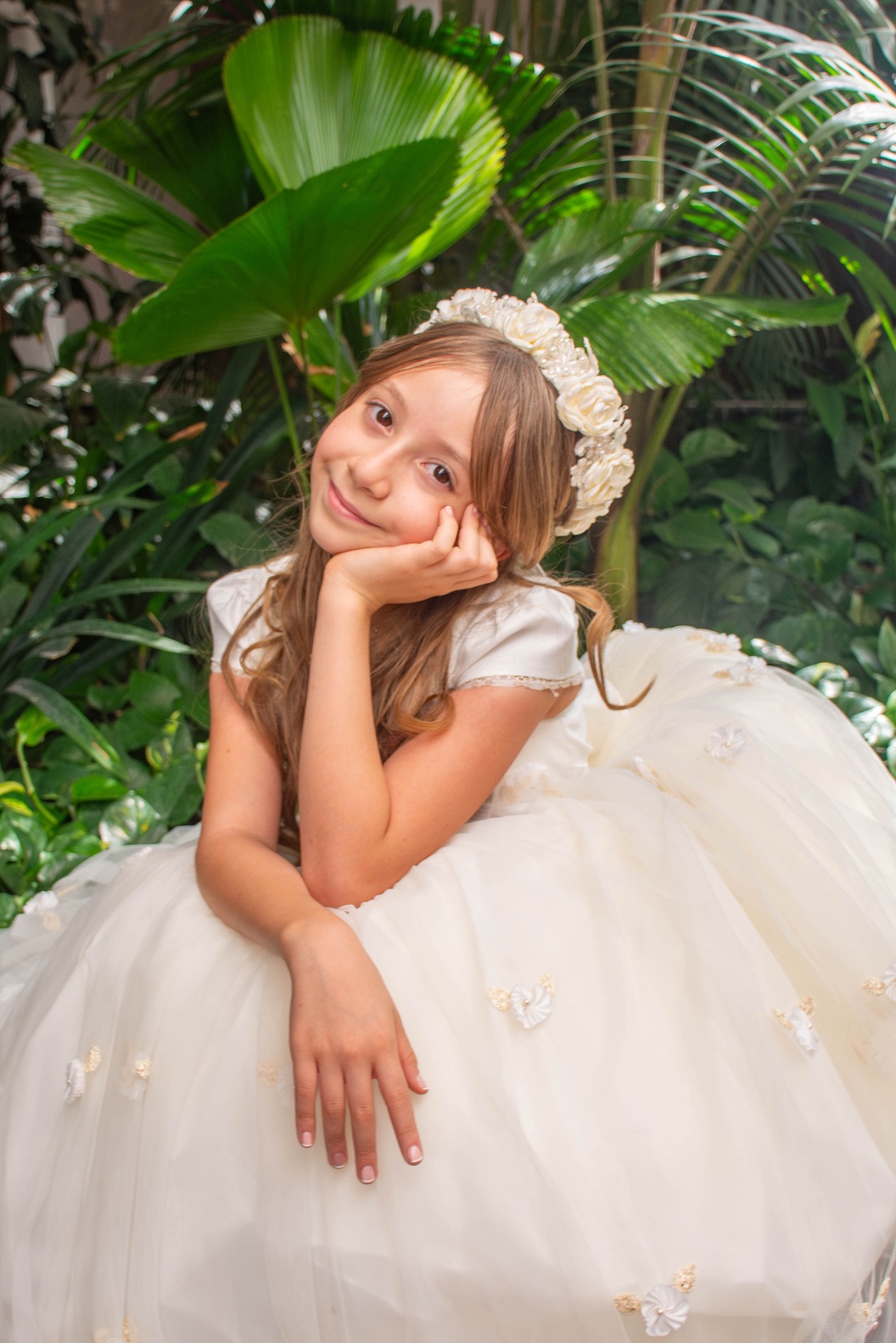 Niña con vestido de primera comunión sonriendo junto a plantas verdes